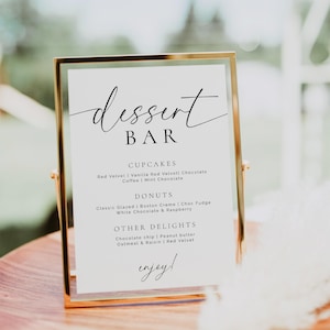 May include: A dessert bar sign in a gold frame. The sign lists cupcakes, donuts, and other delights. Cupcake flavors include Red Velvet and Chocolate. Donut flavors include Classic Glazed and Boston Creme. The sign says "enjoy!"