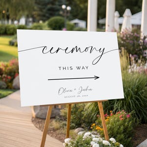 Może przedstawiać: Biały znak na drewnianym sztaludze z napisem "ceremony" w eleganckiej czcionce. Poniżej napis "THIS WAY" ze strzałką w prawo. Wyświetlane są również imiona "Olivia + Joshua" i data. Znak otoczony jest zielenią.