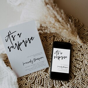 May include: A white card with black text that reads "it's a surprise" and "Kennedy Thompson" with details for a 30th birthday party. The card is on a woven placemat with a dried pampas grass plume.