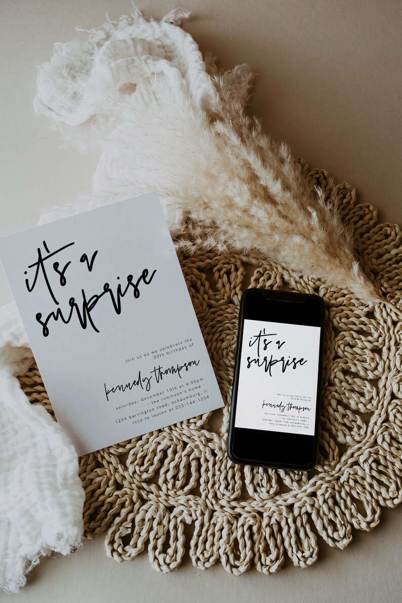 May include: A white card with black text that reads "it's a surprise" and "Kennedy Thompson" with details for a 30th birthday party. The card is on a woven placemat with a dried pampas grass plume.