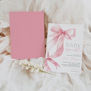 Pu&ograve; includere: Un invito per un baby shower con un design a fiocco rosa e le parole "BABY sprinkle". Una carta rosa con linee verticali &egrave; accanto all'invito. Fiori bianchi in basso.