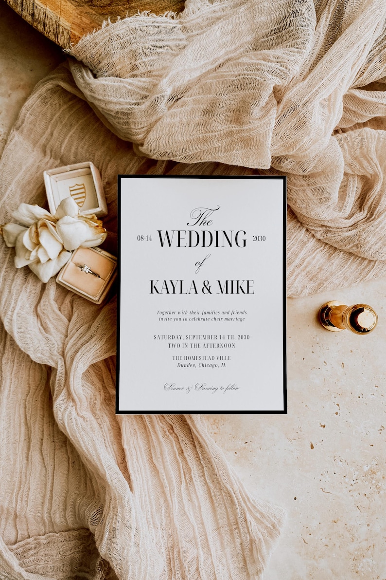 May include: A wedding invitation with the text "The Wedding of Kayla & Mike" in elegant script, set against a neutral backdrop. The invitation is framed in black, with a ring box, flowers, and a gold-toned object nearby.