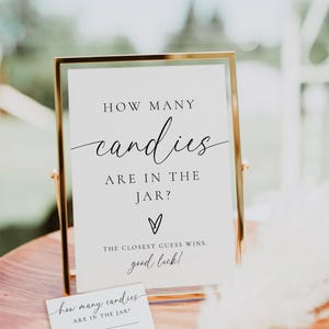 May include: A gold-framed sign with the text "HOW MANY candies ARE IN THE JAR?" and "THE CLOSEST GUESS WINS. good luck!" A small card with fill-in-the-blank lines is also present. The sign is on a wooden surface.