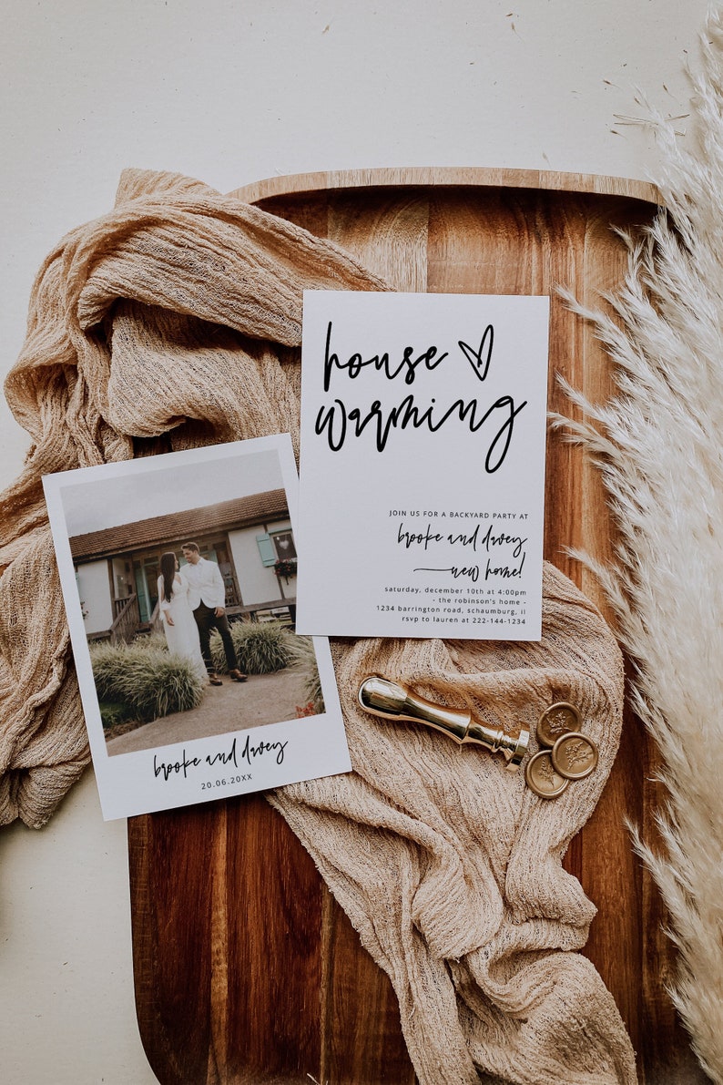May include: A housewarming invitation with a photo of a couple in front of a house. The invitation is on a wooden surface with a beige fabric and a gold wax seal. The text on the invitation reads "house warming" and "brooke and dewey - new home!"
