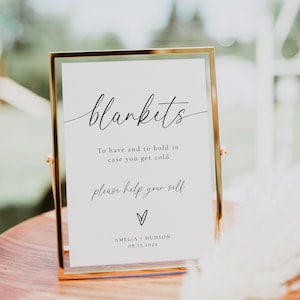 May include: A framed sign with the word "blankets" in elegant script. The sign includes the text "To have and to hold in case you get cold, please help yourself" and the names Amelia and Hudson with a date. The frame is gold.