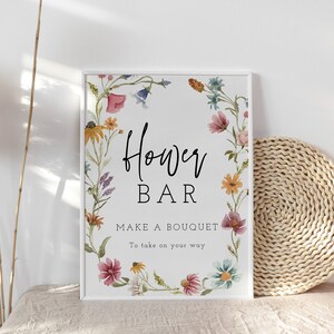 May include: A white sign with a floral border and the text "Flower Bar" in black. Below the text is the phrase "Make a bouquet to take on your way".