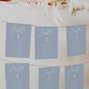 Pode incluir: Seis cart&otilde;es azuis claros com texto branco, cada um com a etiqueta "mesa" seguida de um n&uacute;mero de 1 a 6. Cada cart&atilde;o lista um grupo de nomes para um mapa de lugares de casamento.