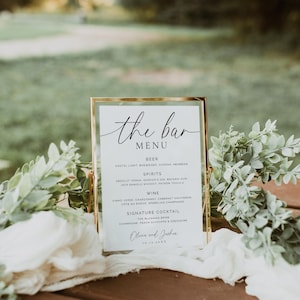 May include: A gold-framed bar menu with the words "the bar MENU" in elegant script. The menu lists beer, spirits, wine, and a signature cocktail. The names Olivia and Joshua are at the bottom, along with a date. Greenery and white fabric surround the menu.