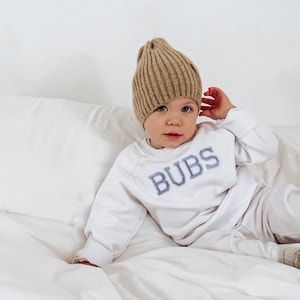 May include: A child wearing a beige ribbed knit beanie, a white sweatshirt with "BUBS" embroidered in blue, and matching white pants. The child is also wearing gray sneakers and is lying on a white bed.