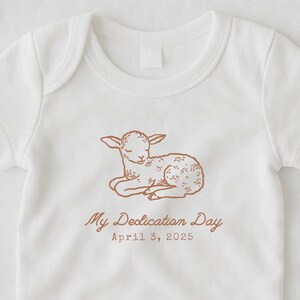 May include: White baby onesie with a brown illustration of a lamb and the text "My Dedication Day April 3, 2025". The onesie has short sleeves and a snap closure at the bottom. The lamb illustration has floral details.