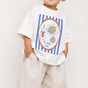May include: A toddler wearing a white t-shirt with a blue and brown graphic design and the words "la dolce vita". The shirt is paired with beige linen pants. The child is smiling and looking to the side.