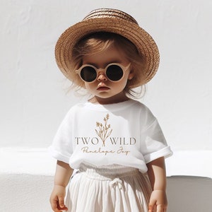 Puede incluir: Un ni&ntilde;o peque&ntilde;o con una camiseta blanca con el texto "TWO WILD Penelope Joy" y un dise&ntilde;o floral. El ni&ntilde;o tambi&eacute;n lleva un sombrero de paja, gafas de sol, una falda blanca y zapatillas blancas.