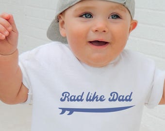 May include: A white t-shirt with a blue graphic of a surfboard and the text "Rad like Dad". A young child is wearing the shirt and a gray baseball cap.