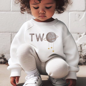 May include: A white sweatshirt with the word "TWO" and a gray planet with gold stars and a ring around it. A young child is wearing the sweatshirt and is squatting down.