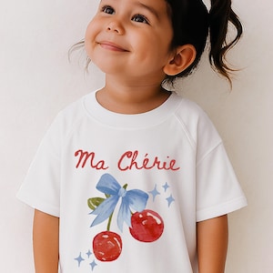 May include: White short-sleeved t-shirt featuring the text "Ma Chérie" in red script above a watercolor illustration of two red cherries with a blue bow and sparkling stars. The shirt is worn by a young child.