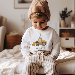 May include: A white long-sleeved shirt with a yellow construction vehicle wearing a blue party hat and the number 7 on it. The shirt has the name Wyatt printed on it.