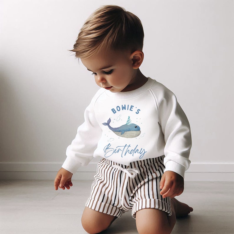 May include: White sweatshirt featuring a blue whale wearing a party hat and the text "BOWIE'S Birthday". A young child is wearing the sweatshirt and looking down.