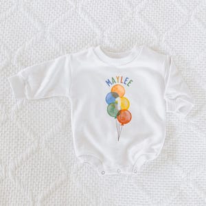 May include: White long-sleeved baby bodysuit with a colorful balloon design. The balloons are in shades of blue, green, orange, and yellow, with the name "MAYLEE" above them. A number "1" is on one of the balloons.