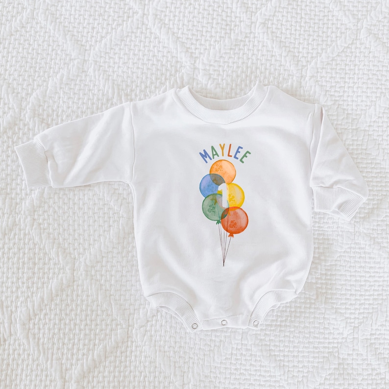 May include: White long-sleeved baby bodysuit with a colorful balloon design. The balloons are in shades of blue, green, orange, and yellow, with the name "MAYLEE" above them. A number "1" is on one of the balloons.