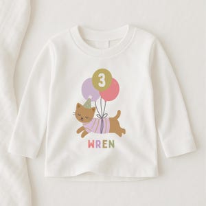 May include: White long-sleeve shirt featuring a cartoon cat with a party hat and balloons. The cat is wearing a pink and purple striped shirt. The number 3 is on the balloons. The name "WREN" is printed below the cat in rainbow colors.