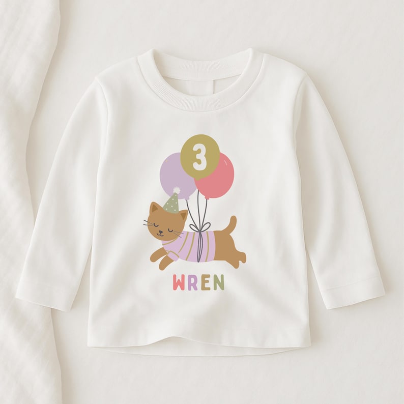 May include: White long-sleeve shirt featuring a cartoon cat with a party hat and balloons. The cat is wearing a pink and purple striped shirt. The number 3 is on the balloons. The name "WREN" is printed below the cat in rainbow colors.