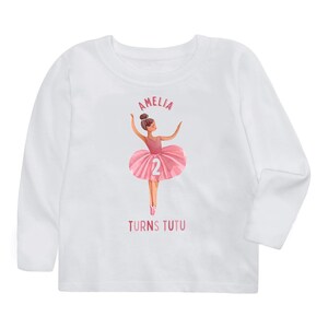 May include: White long-sleeved t-shirt with a pink ballerina illustration and the text "Amelia Turns Tutu" and the number "2" on the ballerina's tutu.
