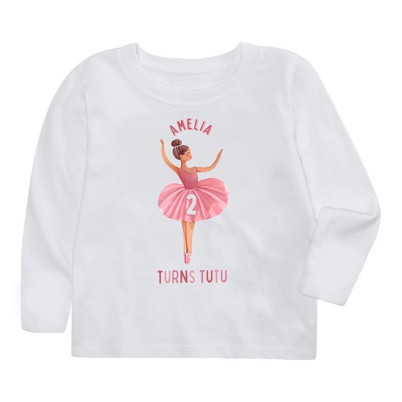 May include: White long-sleeved t-shirt with a pink ballerina illustration and the text "Amelia Turns Tutu" and the number "2" on the ballerina's tutu.