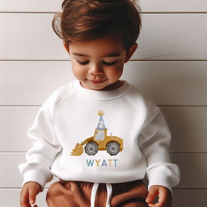 May include: White sweatshirt with a yellow construction vehicle wearing a blue party hat with the number 2 on it. The text "WYATT" is printed below the vehicle in blue, yellow, and orange.