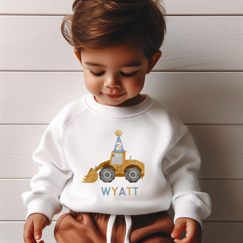May include: White sweatshirt with a yellow construction vehicle wearing a blue party hat with the number 2 on it. The text "WYATT" is printed below the vehicle in blue, yellow, and orange.