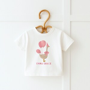 May include: White short-sleeved t-shirt with a pink and brown goose wearing a party hat and holding pink balloons. The text "Emma Grace" is printed below the goose.