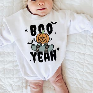 Op de afbeelding: Witte baby-onesie met een Halloween-thema. Het ontwerp bevat de woorden "BOO YEAH" in zwart, een cartoon pompoen op een skateboard, vleermuizen en sterren. De onesie is gemaakt van een zacht materiaal.