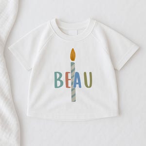 Pode incluir: T-shirt branca de manga curta com um gr&aacute;fico de vela colorida e o nome "BEAU" em letras azuis, vermelhas e verdes. A vela tem uma chama amarela e um corpo listrado azul e branco. Artigo de vestu&aacute;rio infantil.