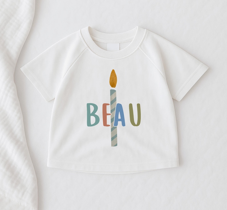 Pode incluir: T-shirt branca de manga curta com um gr&aacute;fico de vela colorida e o nome "BEAU" em letras azuis, vermelhas e verdes. A vela tem uma chama amarela e um corpo listrado azul e branco. Artigo de vestu&aacute;rio infantil.