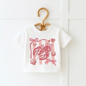 May include: White t-shirt with a pink lobster graphic, seashells, bows, and hearts. The design includes pink vertical stripes and the number "11". The shirt is hanging on a wooden hanger.