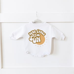 May include: White long-sleeved baby onesie with a gold graphic that says "Here Comes The Sun" with a stylised sun graphic.