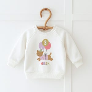 May include: White baby sweatshirt with a cat graphic holding balloons. The cat wears a party hat and a striped shirt. The number "3" is on the balloons. The name "WREN" is printed below the cat in pastel colors.