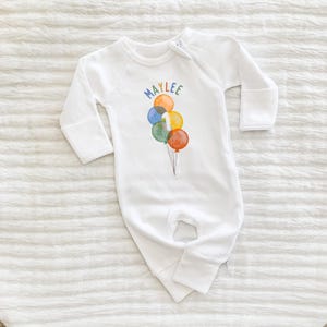 May include: White baby sleeper with long sleeves and a zipper closure. The front features a colorful watercolor design of balloons and the name "MAYLEE" in rainbow letters. A number "1" is on one of the balloons.