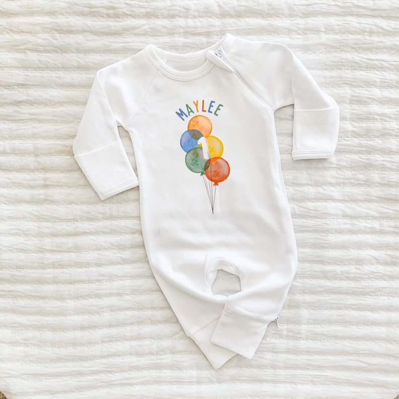 May include: White baby sleeper with long sleeves and a zipper closure. The front features a colorful watercolor design of balloons and the name "MAYLEE" in rainbow letters. A number "1" is on one of the balloons.