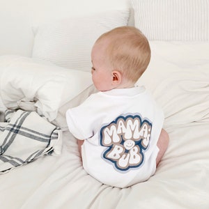 May include: A white baby onesie with a blue, brown, and white graphic that says "Mama's Bub" with a smiley face in the middle of the word "Bub".