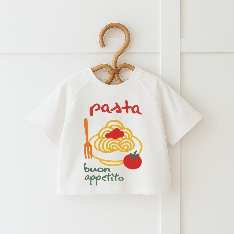Op de afbeelding: Wit T-shirt met korte mouwen met een pasta-thema. Het ontwerp bevat het woord "pasta" in het rood, een bord spaghetti met tomatensaus, een vork, een tomaat en de woorden "buon appetito" in het groen.