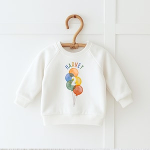 May include: White crewneck sweatshirt with the name "HARVEY" in rainbow letters above a cluster of watercolor balloons. The balloons are in shades of orange, blue, yellow, green, and red. The number "2" is on one of the balloons.