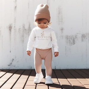Puede incluir: Un ni&ntilde;o peque&ntilde;o con una camiseta blanca de manga larga con el texto "TWO WILD Penelope Joy" y una boina marr&oacute;n. El ni&ntilde;o lleva pantalones de color beige y zapatillas blancas. El ni&ntilde;o est&aacute; de pie en una plataforma de madera.