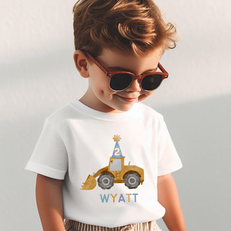 May include: A white t-shirt with a yellow construction vehicle wearing a blue party hat with the number 2 on it. The text "WYATT" is printed below the vehicle.