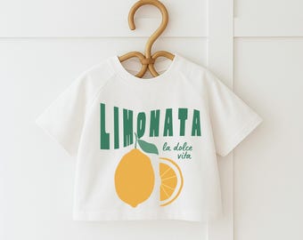 May include: White short-sleeved t-shirt with a lemon graphic and the text "LIMONATA la dolce vita" in green. The graphic includes a whole lemon and a lemon slice. The shirt is hanging on a wooden hanger.