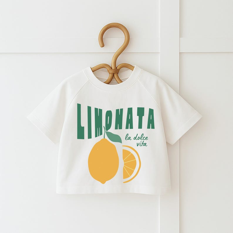 May include: White short-sleeved t-shirt featuring a lemon graphic and the text "LIMONATA la dolce vita" in green. The design includes a whole lemon and a lemon slice. The shirt is hanging on a wooden hanger.