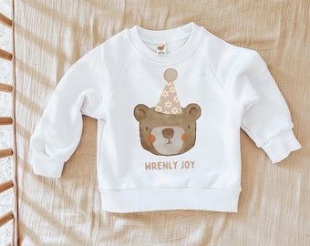 May include: White sweatshirt with a brown bear wearing a pink and white party hat with white flowers. The text "WRENLY JOY" is printed below the bear.