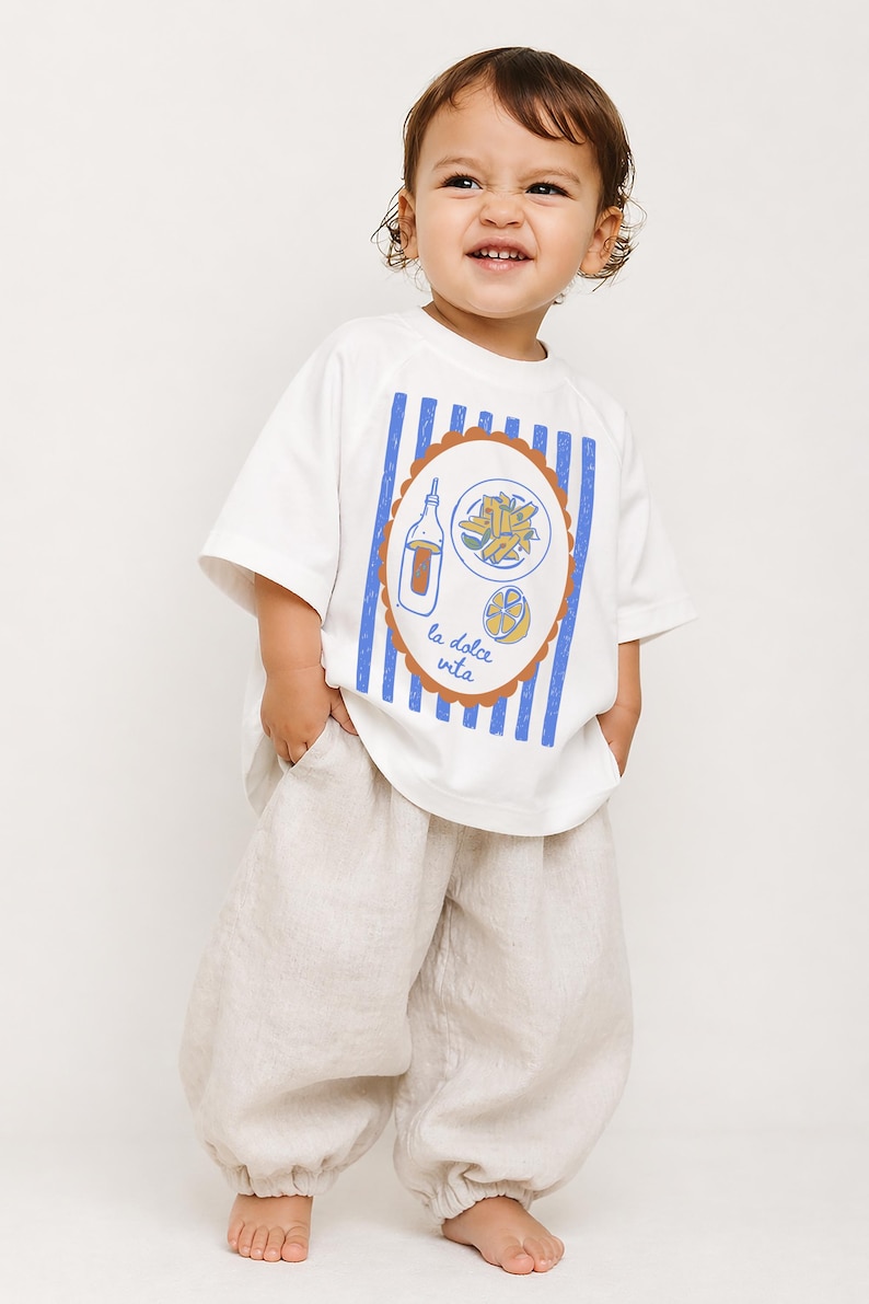 May include: A toddler wearing a white t-shirt with a blue and brown graphic design and the words "la dolce vita". The shirt is paired with beige linen pants. The child is smiling and looking to the side.