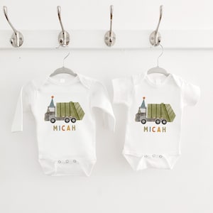 May include: Two white baby onesies with long and short sleeves. Each onesie features a green and brown cartoon garbage truck with a party hat on top. The text "MICAH" is printed below the truck in colorful letters.