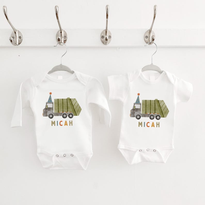 May include: Two white baby onesies with long and short sleeves. Each onesie features a green and brown cartoon garbage truck with a party hat on top. The text "MICAH" is printed below the truck in colorful letters.