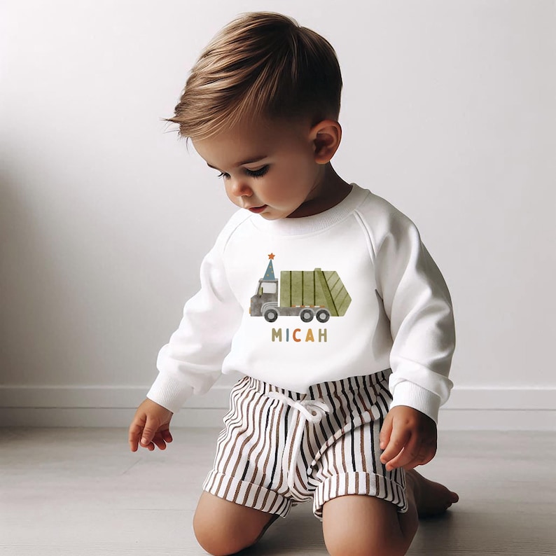 May include: A white sweatshirt with a green garbage truck wearing a party hat and the name Micah printed below. A young child is wearing the sweatshirt and brown and white striped shorts.